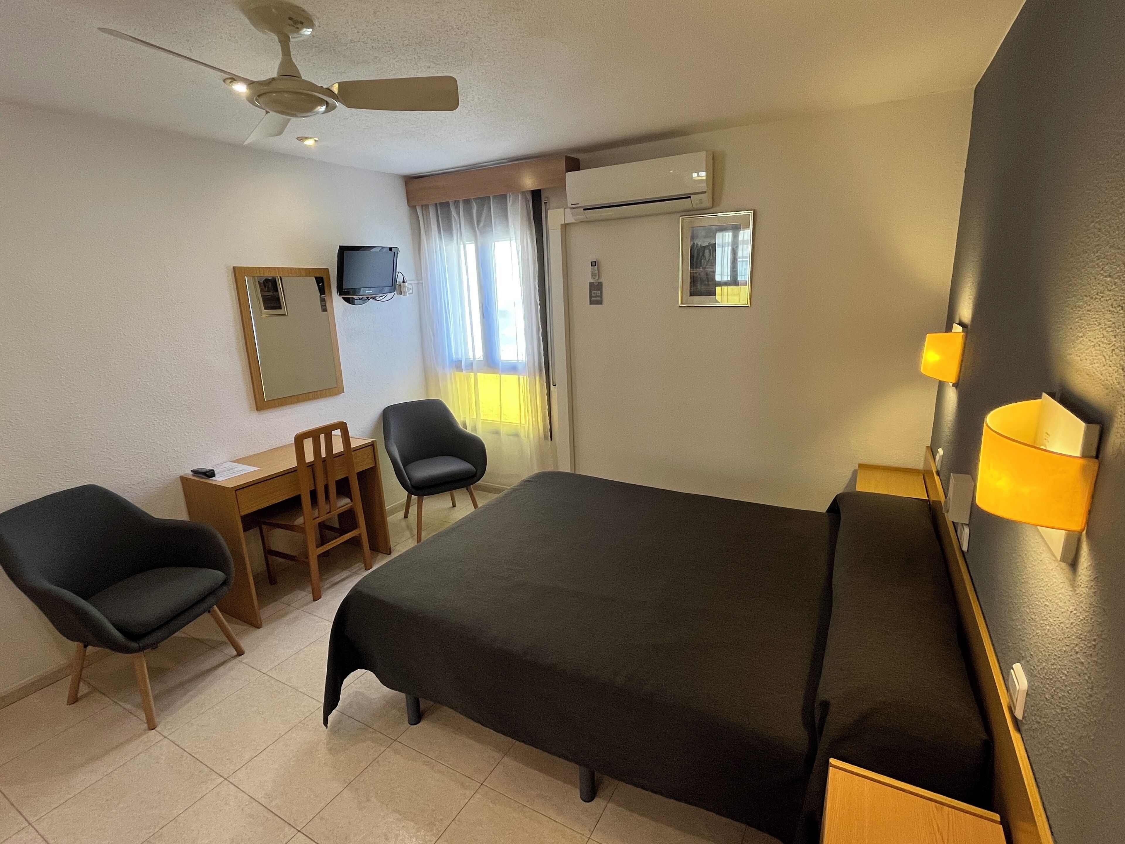 A hotel room with a bed, two chairs, a desk, a television, an air conditioning unit, and a window with curtains.