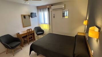 Una habitación de hotel con cama, dos sillas, escritorio, televisor, aire acondicionado y ventana con cortinas.