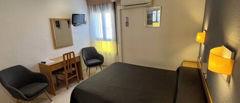 A hotel room with a bed, two chairs, a desk, a television, an air conditioning unit, and a window with curtains.