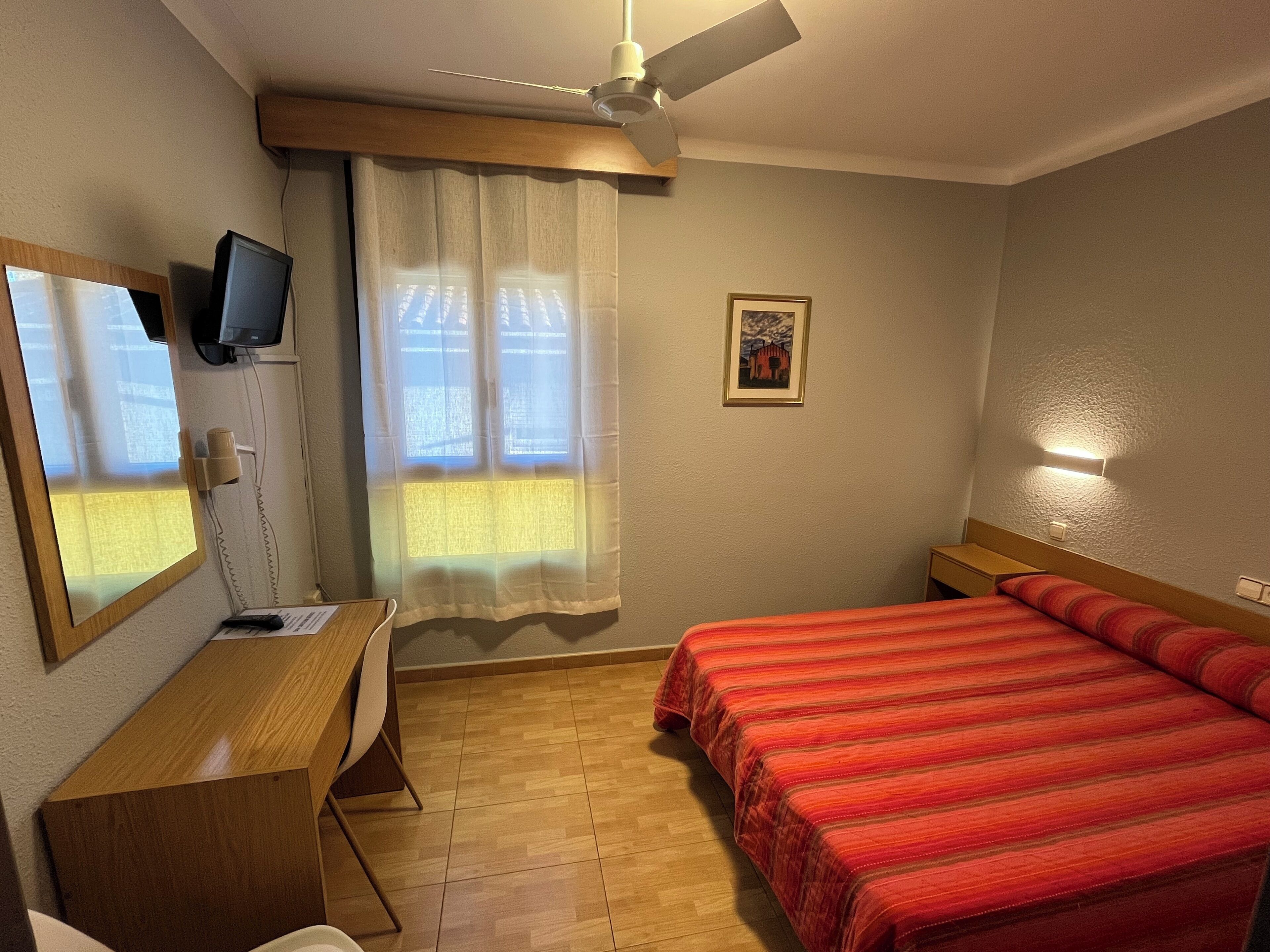 A hotel room with a bed, desk, chair, ceiling fan, window with curtains, and a wall-mounted TV.