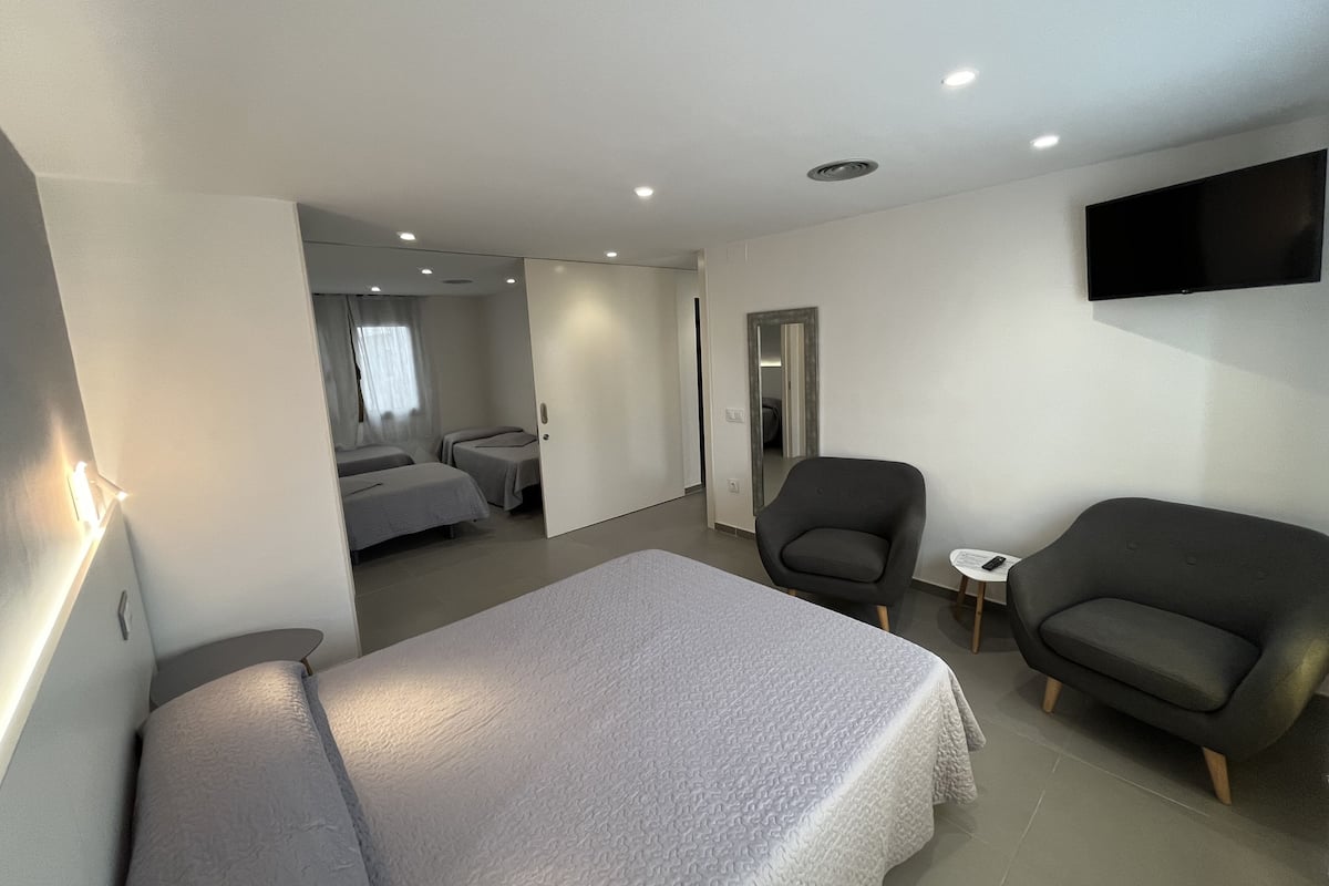 Una habitación de hotel moderna con una cama grande, dos sillones, una mesita, un televisor montado en la pared y un espejo.