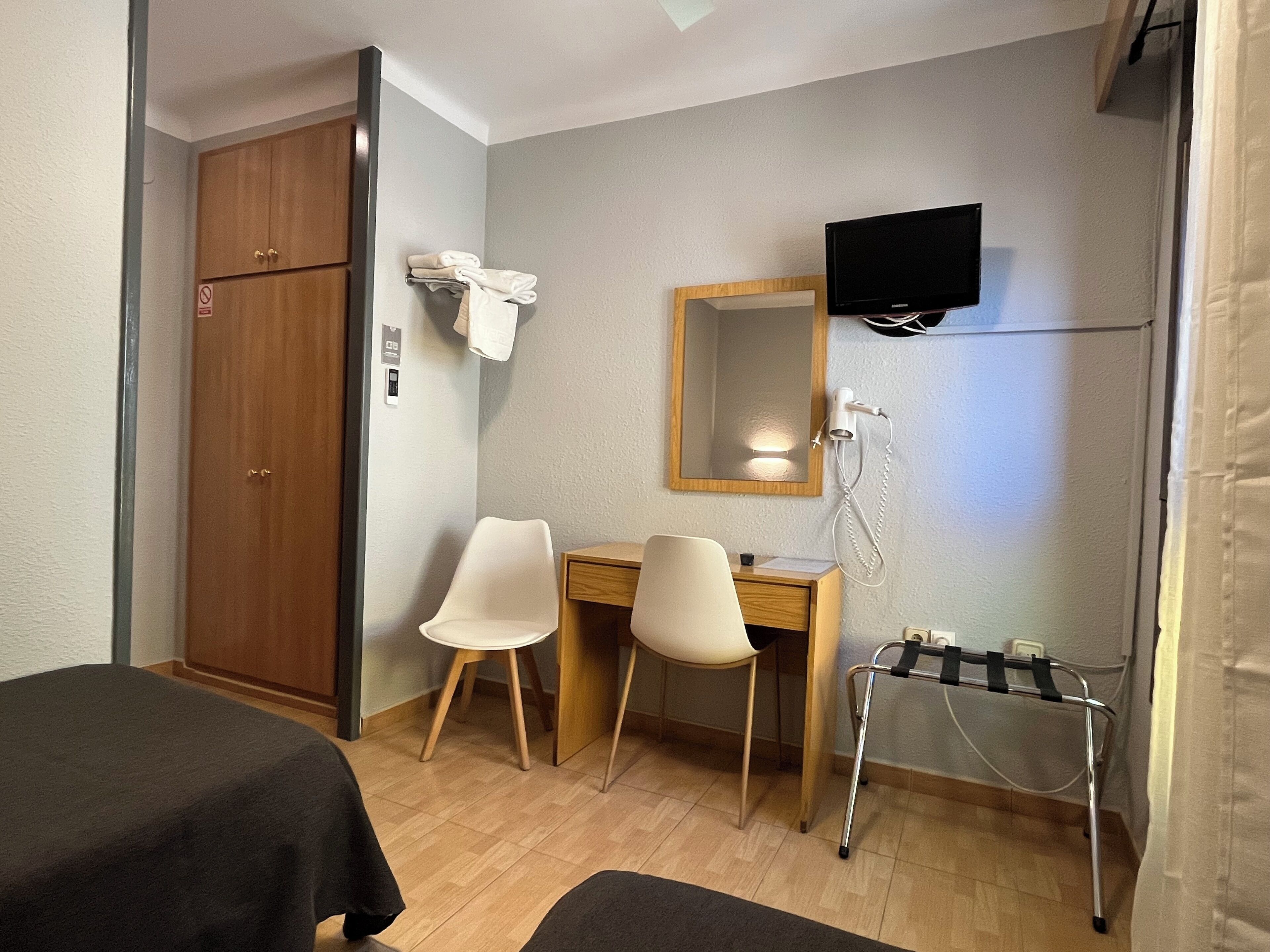 Habitación de hotel con un armario de madera, dos sillas blancas, un escritorio, un televisor montado en la pared y un secador de pelo en un soporte de pared.