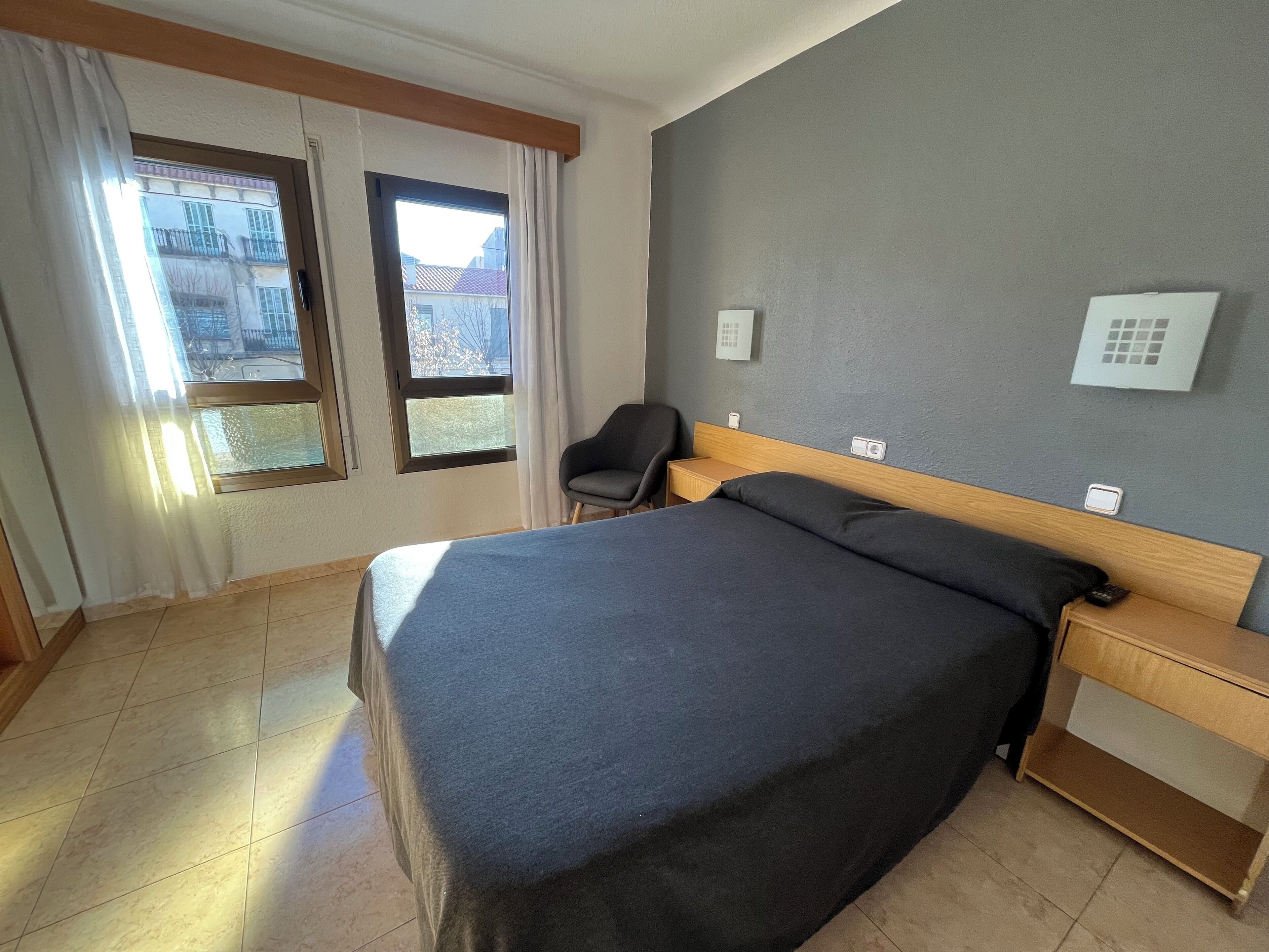 A hotel room with a bed, a chair, a nightstand, a window with curtains, and a thermostat on the wall.