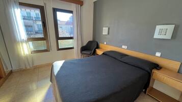 A hotel room with a bed, a chair, a nightstand, a window with curtains, and a thermostat on the wall.