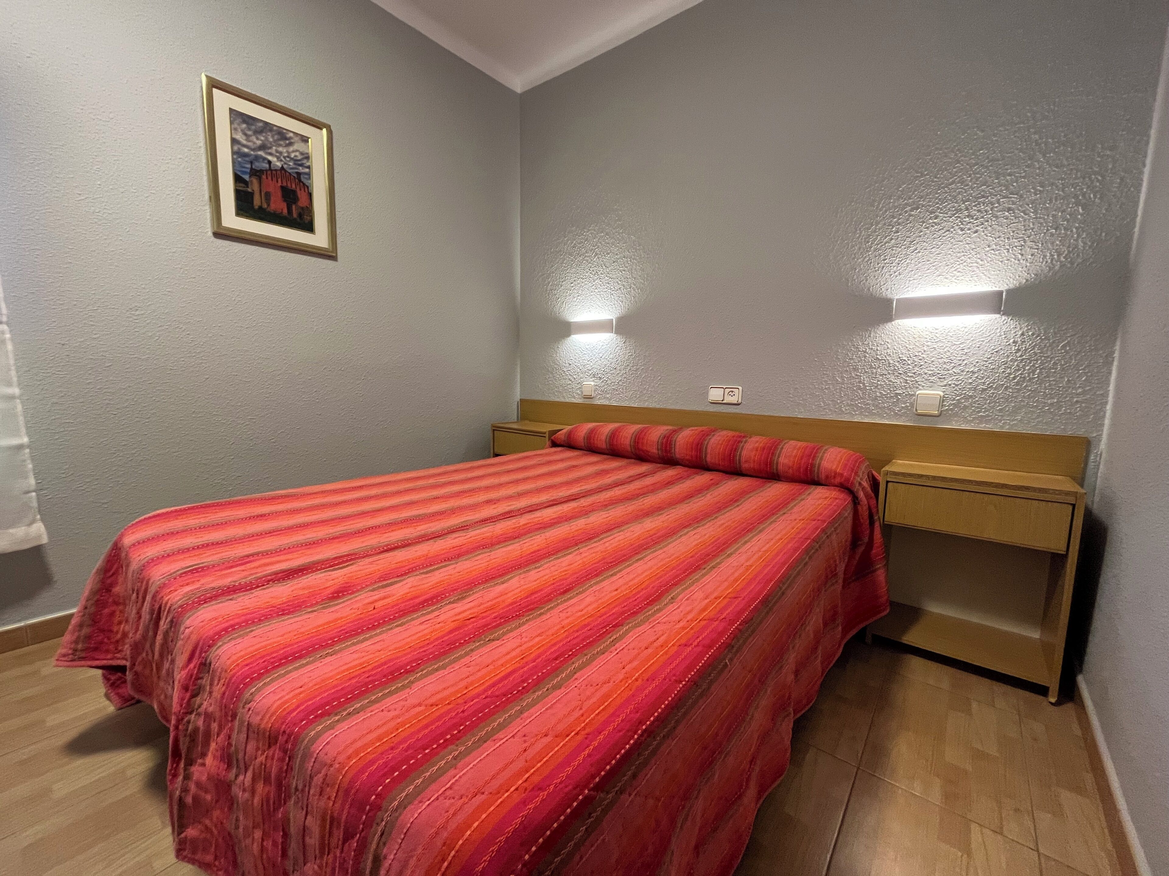 A bedroom with a bed, a nightstand, a wall-mounted light, and a framed picture on the wall.