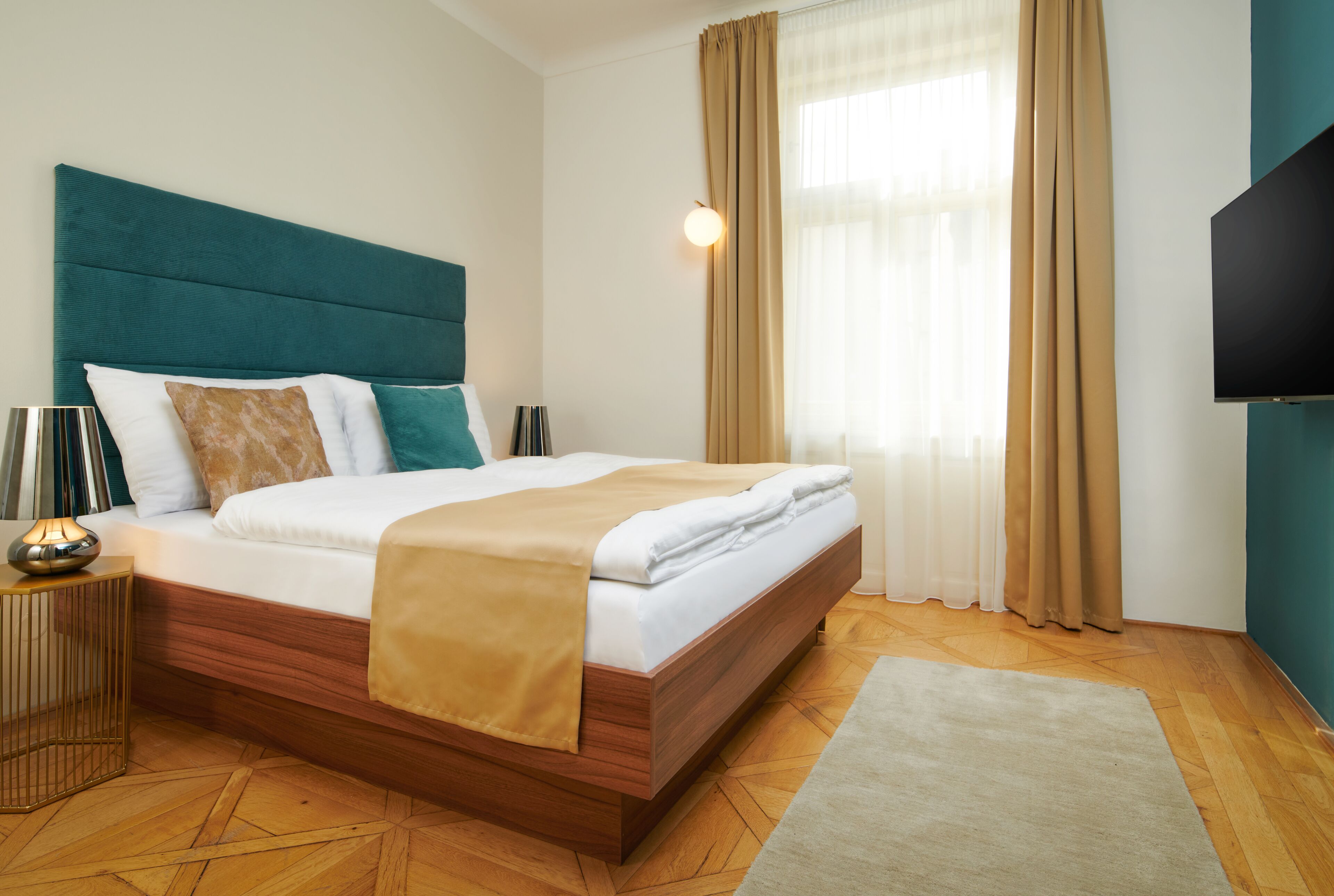 Une chambre d’hôtel moderne avec une tête de lit en bois, un grand lit avec du linge de lit blanc, un plaid beige, une lampe de chevet et un téléviseur à écran plat fixé au mur.