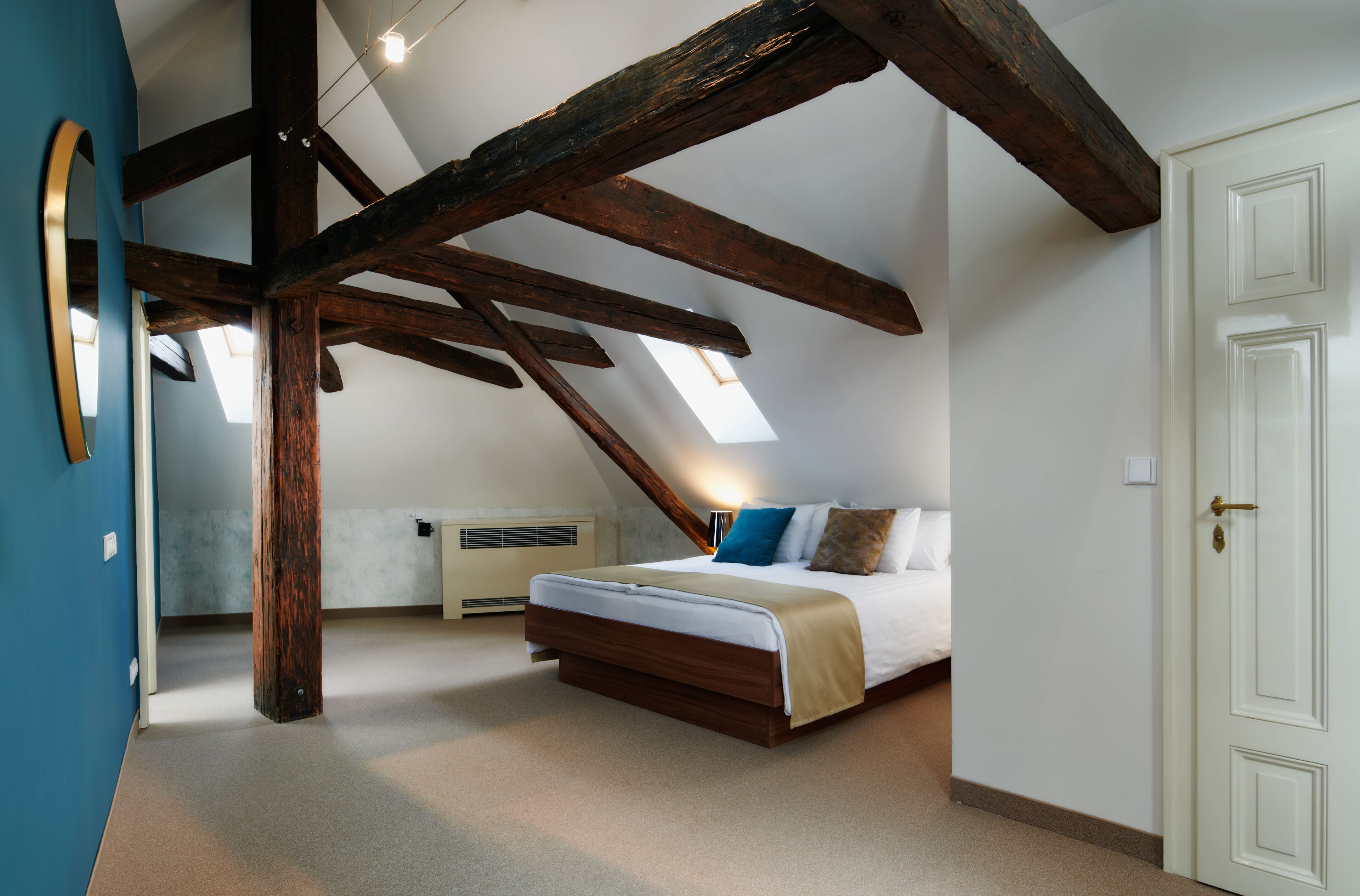 Une chambre avec un plafond en pente, des poutres apparentes, un lit avec un matelas blanc et des oreillers bleus, une porte blanche et un puits de lumière.
