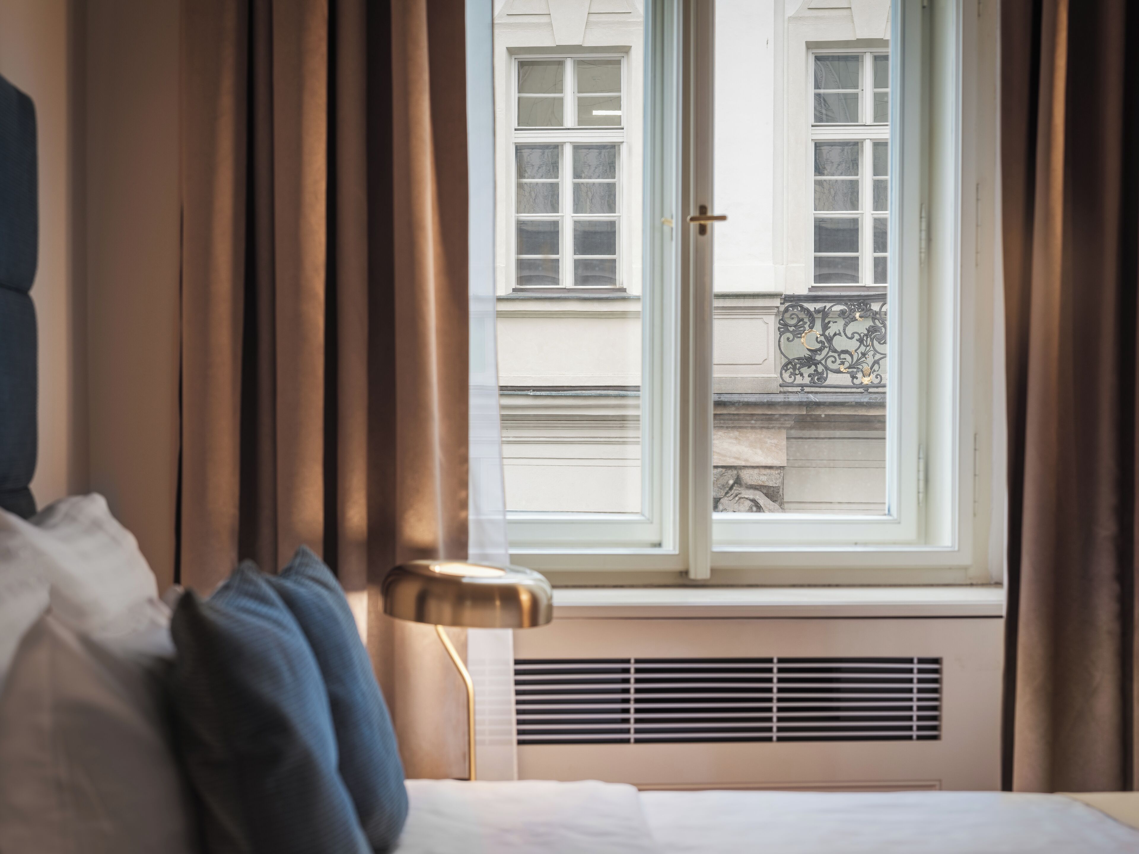 A hotel room with a bed, a window with curtains, a radiator, and a view of a building outside.