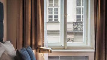 A hotel room with a bed, a window with curtains, a radiator, and a view of a building outside.