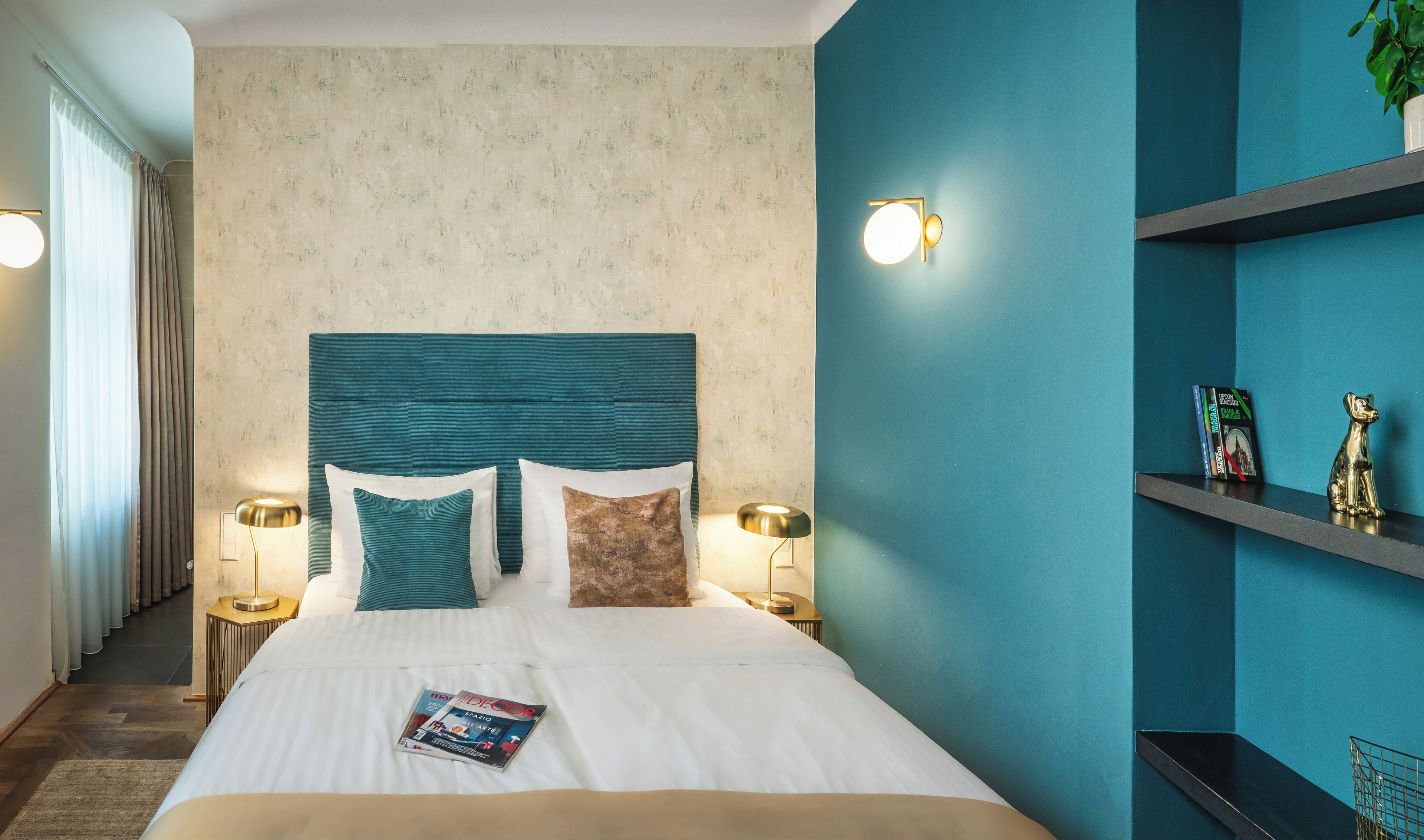 Una cama bien hecha con almohadas, una revista y una mesita de noche con una lámpara. Hay dos apliques de pared y un estante con libros y una pequeña estatua.
