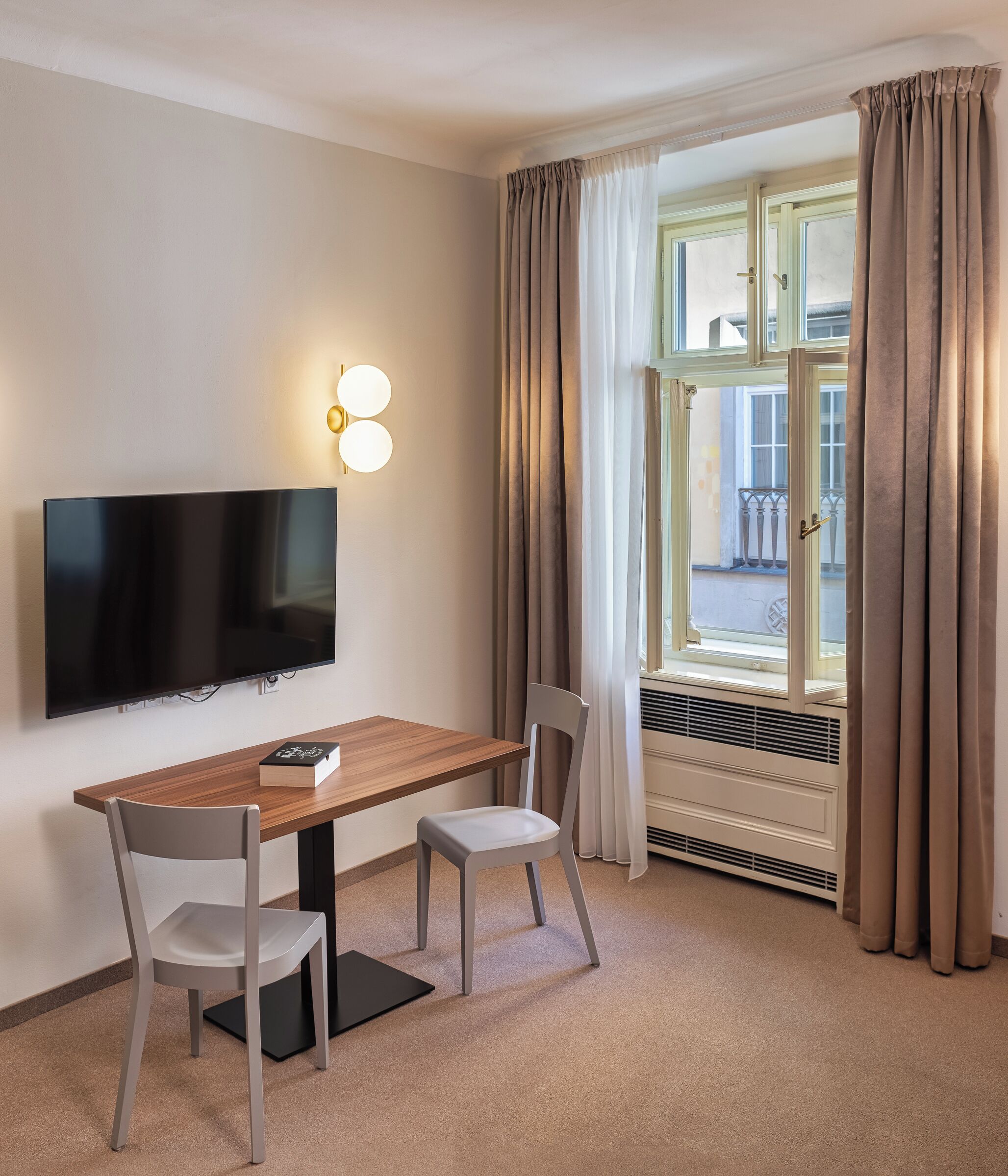 Quarto de hotel com televisão de tela plana na parede, uma mesa de madeira com duas cadeiras, uma janela com cortinas e um radiador.