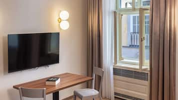 Quarto de hotel com televisão de tela plana na parede, uma mesa de madeira com duas cadeiras, uma janela com cortinas e um radiador.