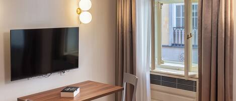 A hotel room with a flat-screen TV mounted on the wall, a wooden table with two chairs, a window with curtains, and a radiator.