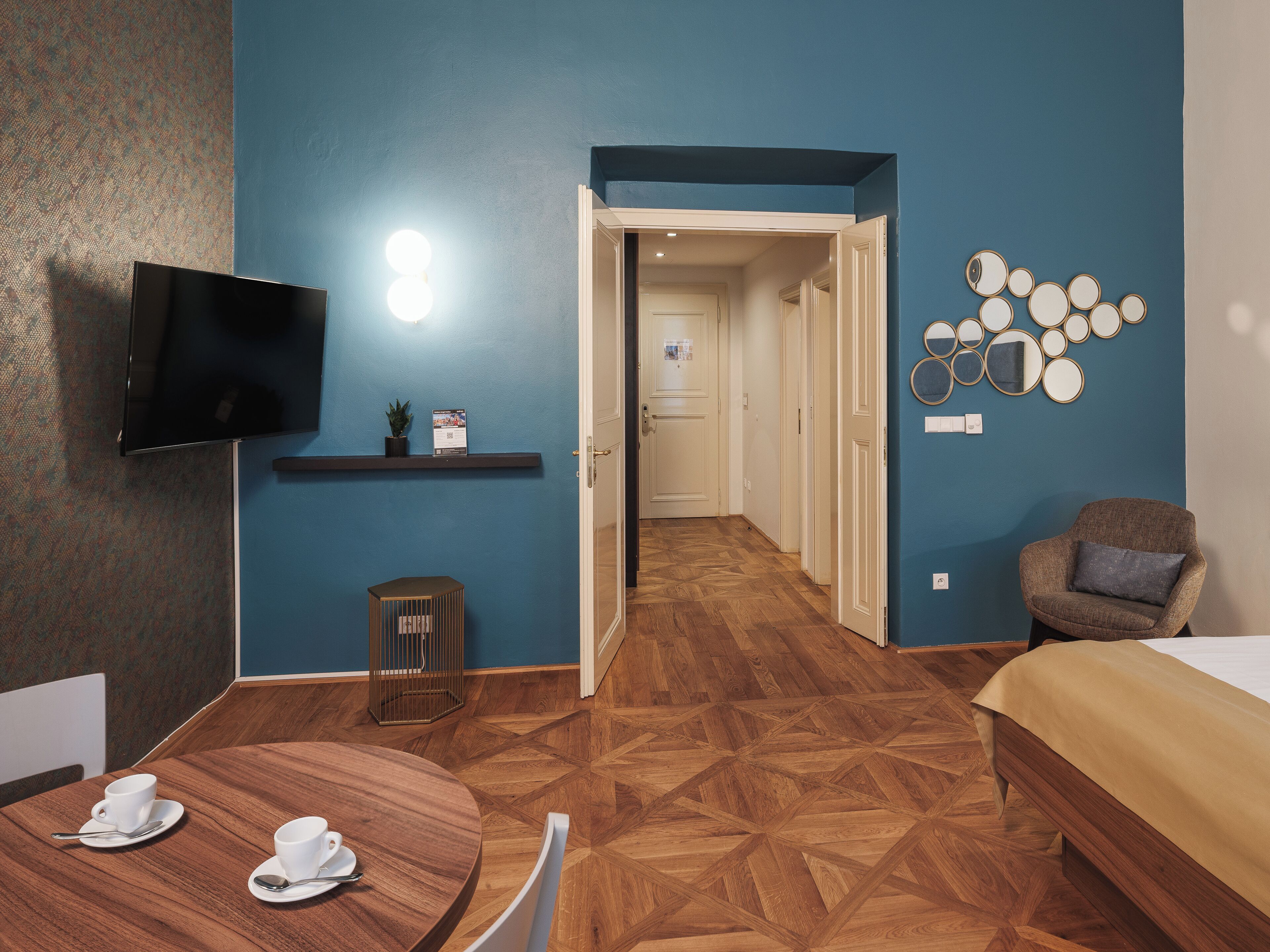 A hotel room with a TV, a small table with cups, a bed, and a hallway leading to another room.