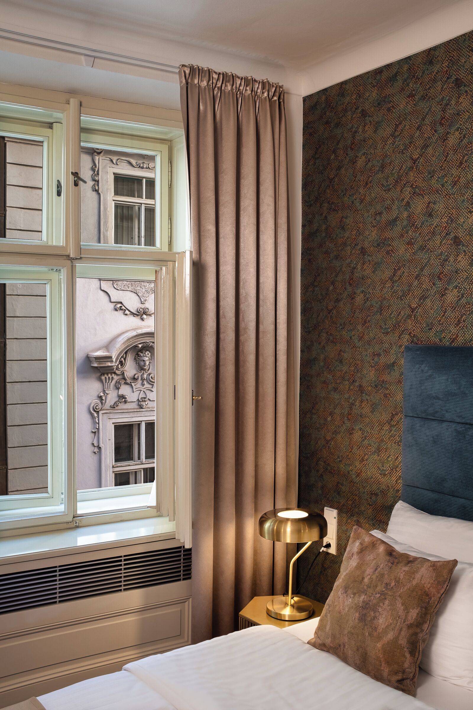 A hotel room with a bed, a nightstand with a lamp, a window with curtains, and a patterned wall.
