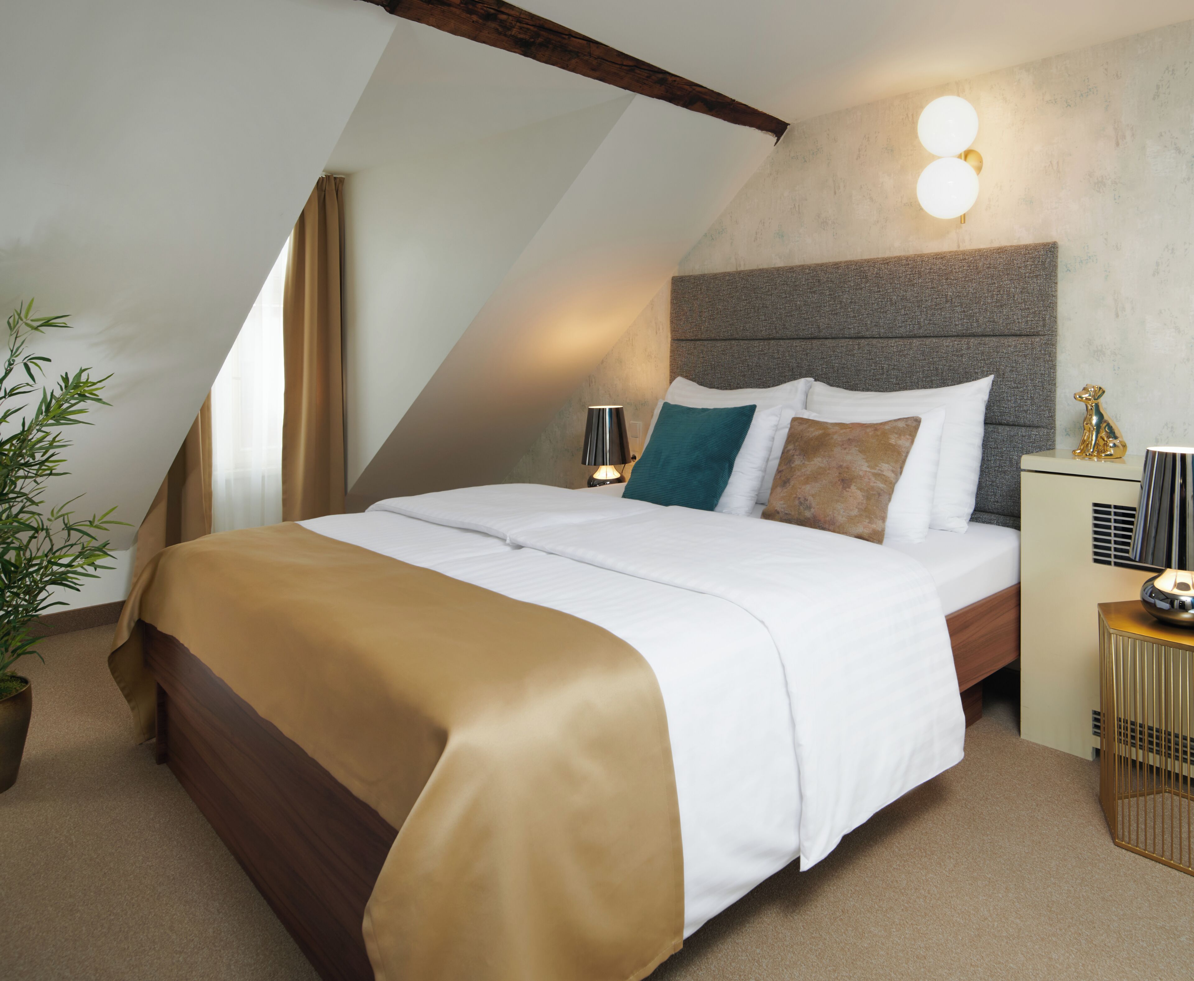 A modern hotel room with a sloped ceiling, a large bed with white and brown bedding, a bedside table with a lamp, and a small plant.