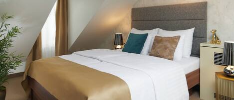 A modern hotel room with a sloped ceiling, a large bed with white and brown bedding, a bedside table with a lamp, and a small plant.