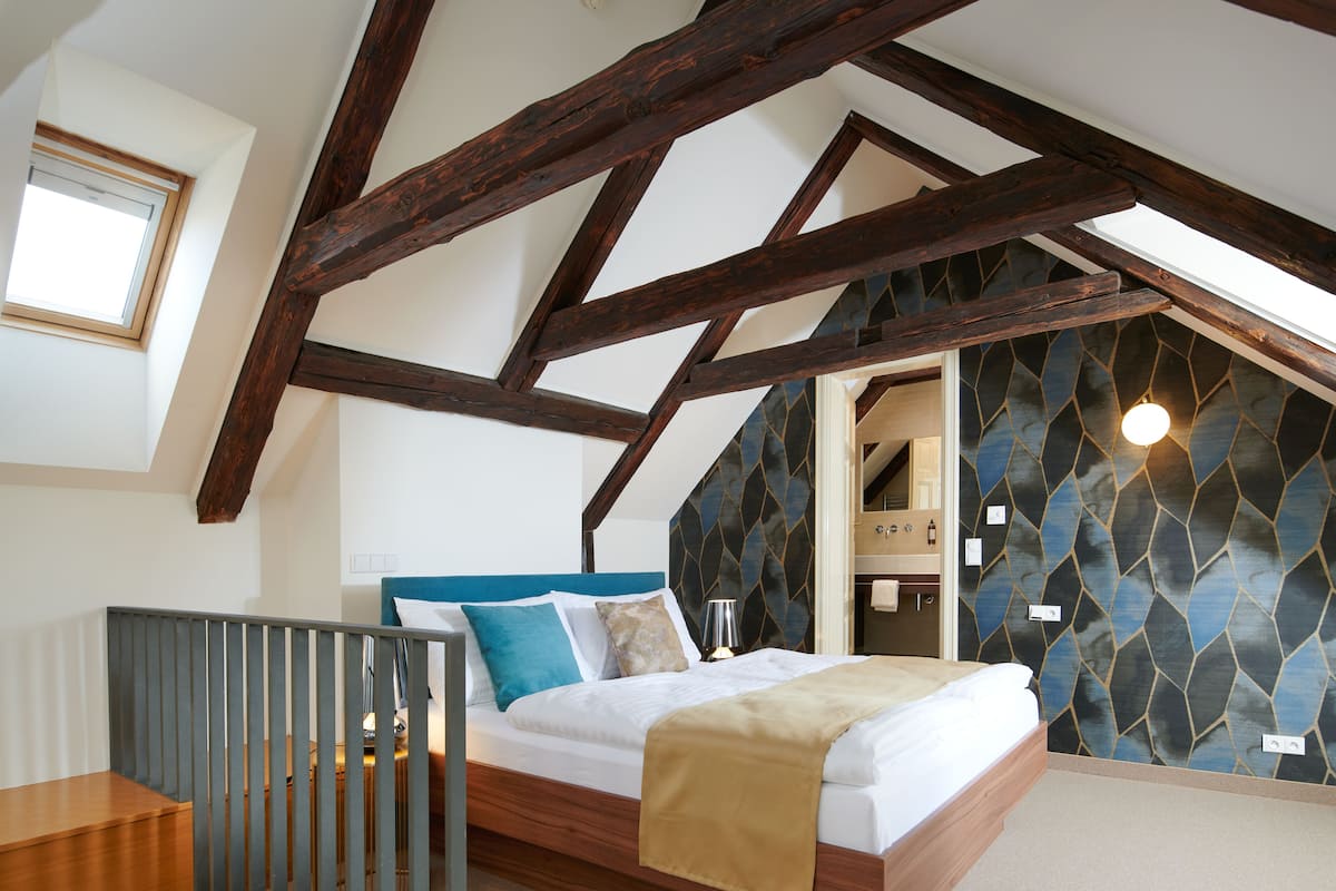 A bedroom with a sloped ceiling, wooden beams, a bed with a blue headboard, a nightstand, and a view into the bathroom.