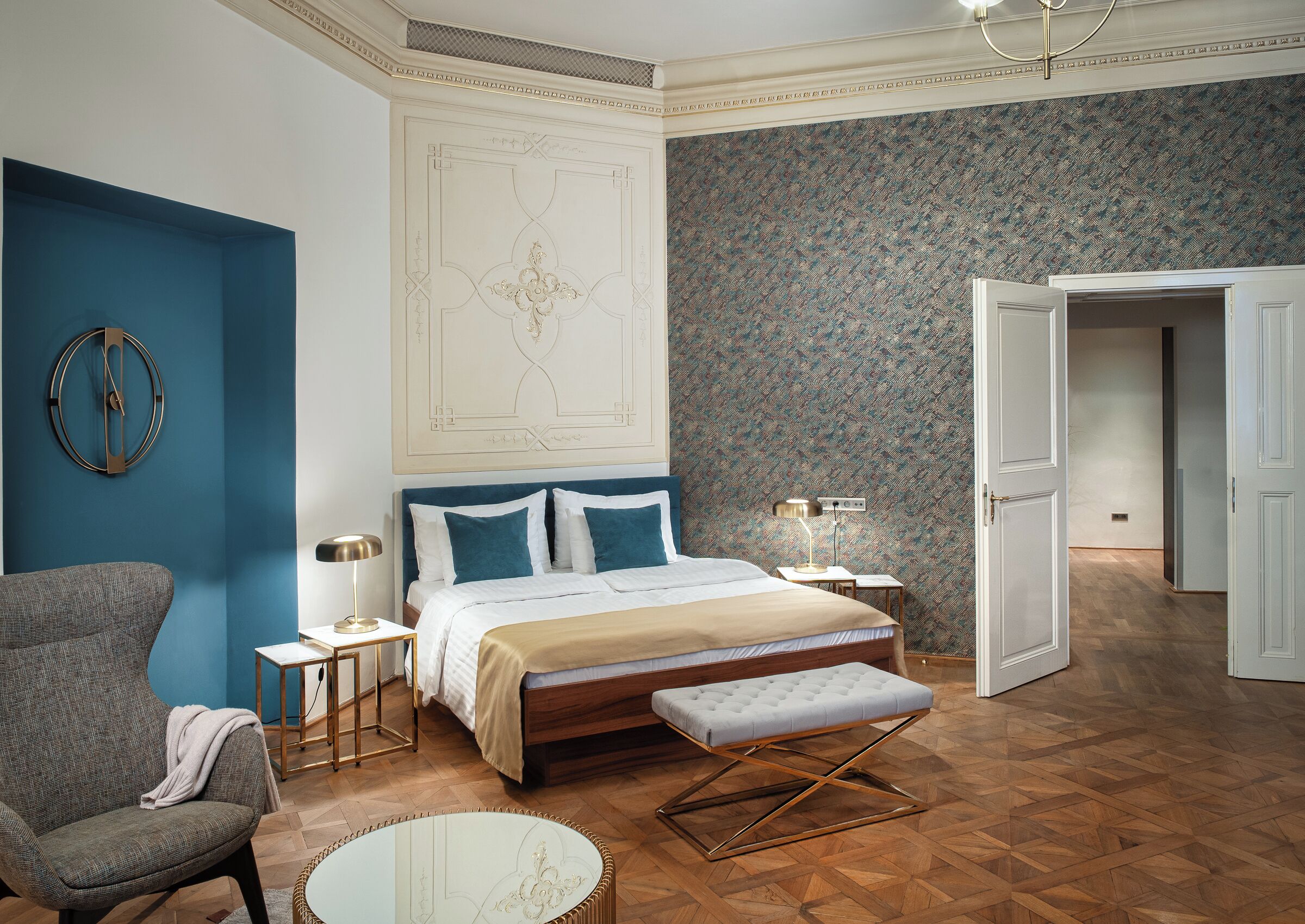A bedroom with a bed, bedside tables, a chair, a round table, and a door leading to another room.