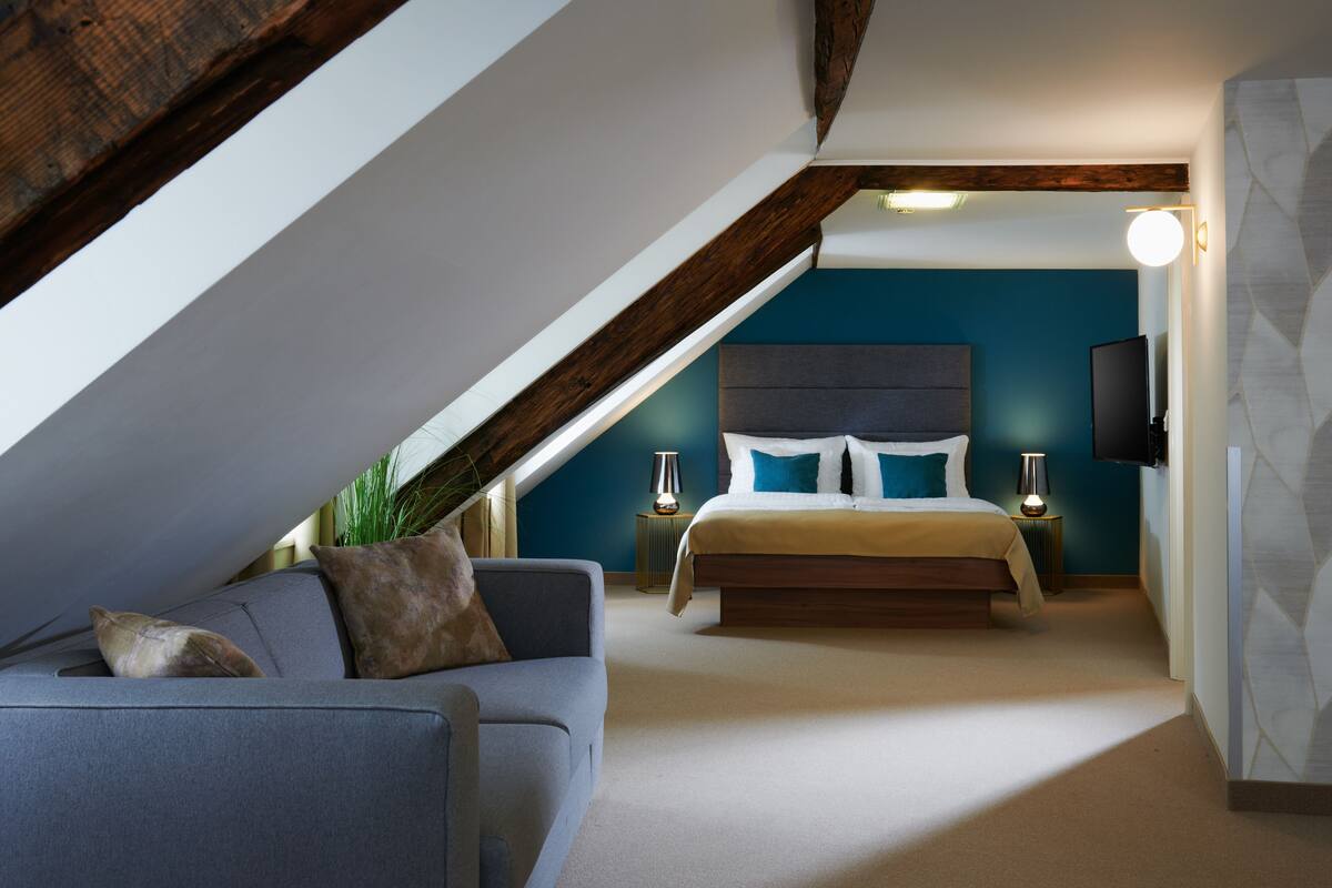 A bedroom with a sloped ceiling, a bed with white and blue bedding, a gray sofa, and two bedside lamps.