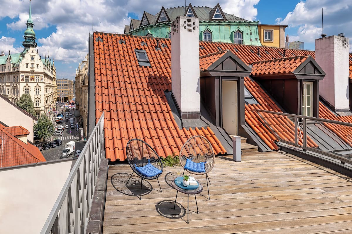 A rooftop terrace with orange-tiled roofs, two wicker chairs, and a small table with a plant.