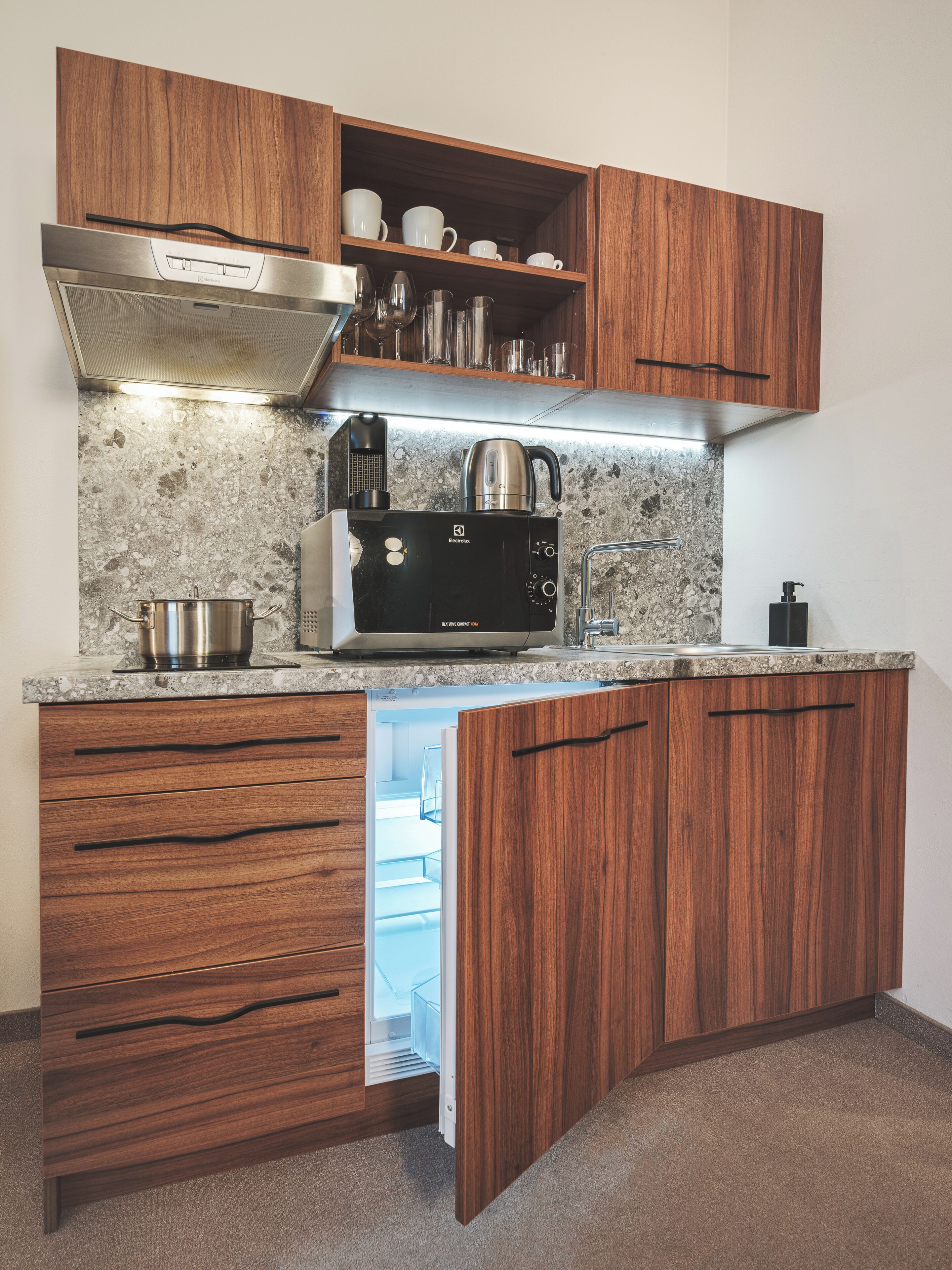 Een kleine keuken met houten kastjes, een ingebouwde magnetron en een roestvrijstalen waterkoker.