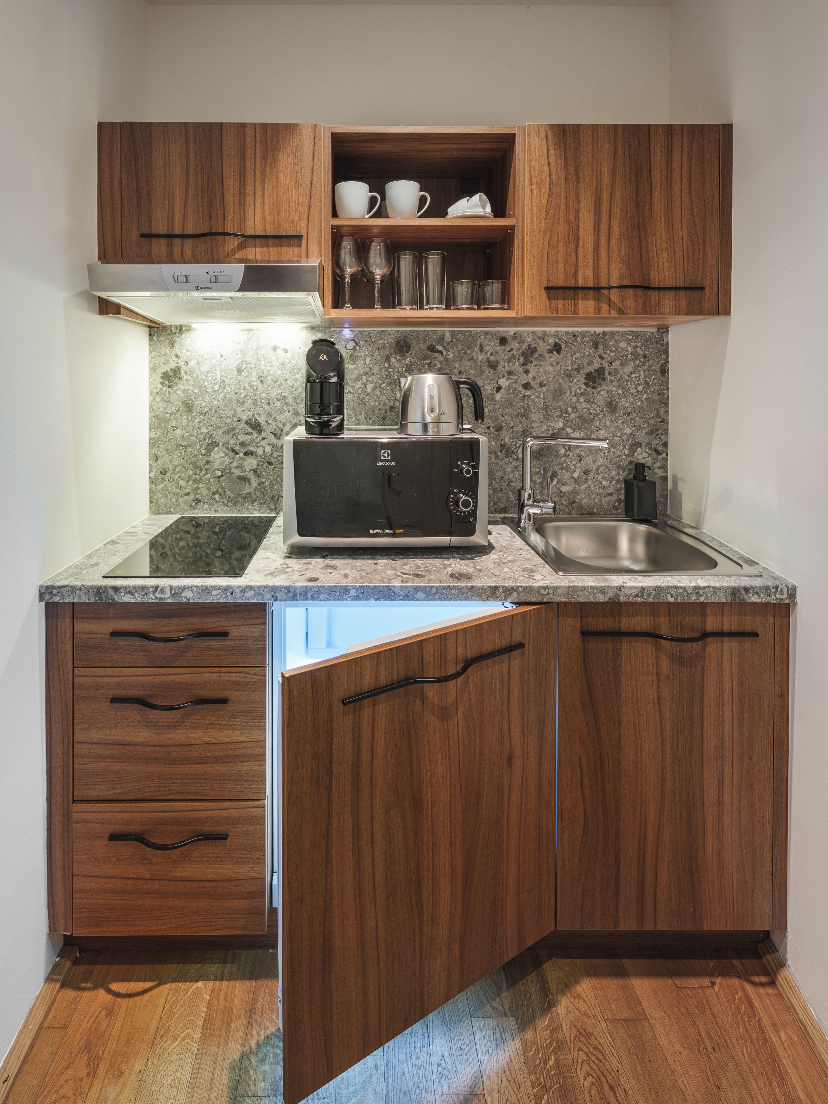A compact kitchen with wooden cabinets, a built-in coffee maker, a kettle, and a stainless steel sink.