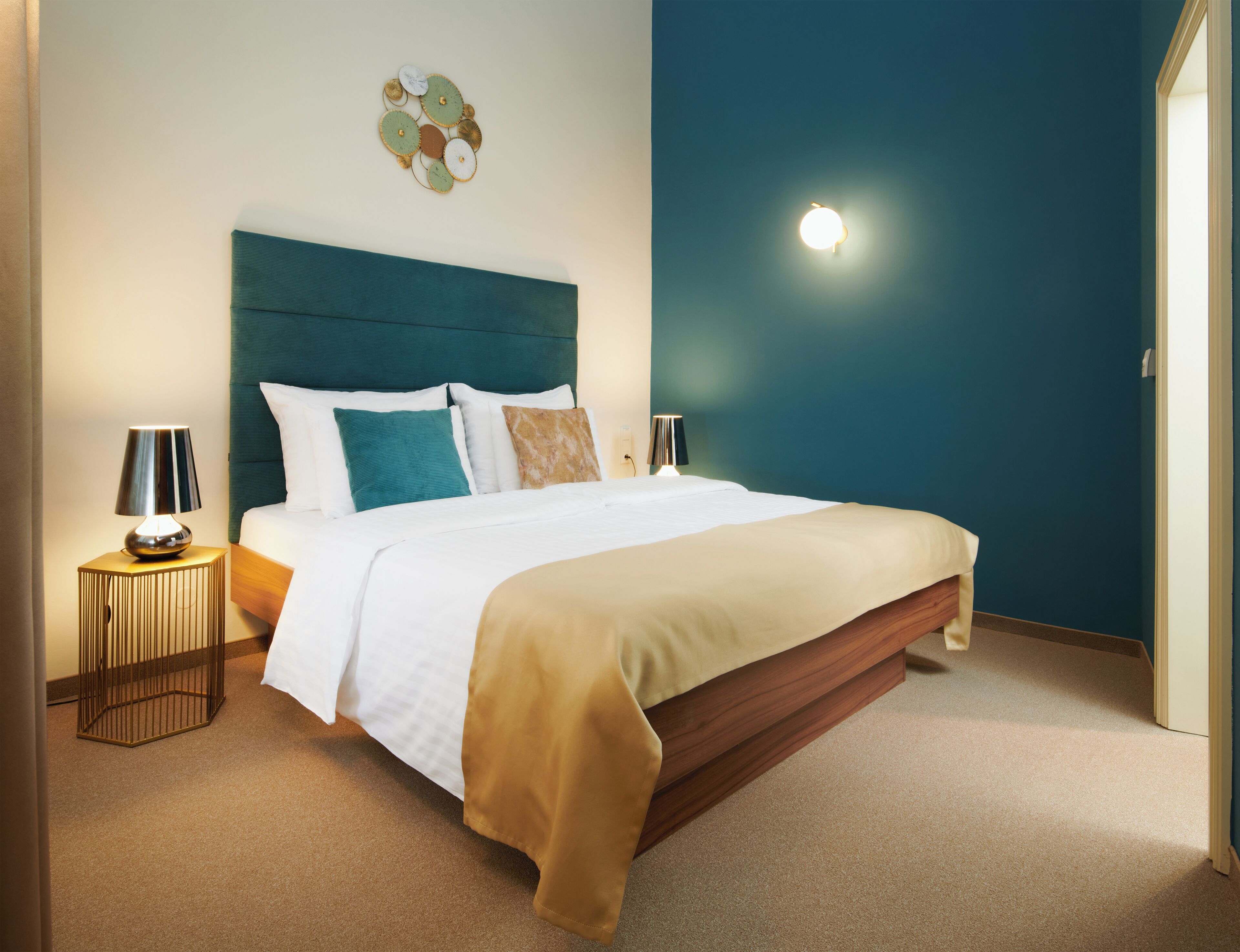 Un lit bien fait, avec une tête de lit bleue, des oreillers blancs et turquoise, et un plaid beige. Il y a deux tables de chevet avec des lampes, et une œuvre d’art murale au-dessus du lit.