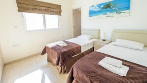 A hotel room with two beds, a window with a brown valance, a painting of a beach scene, and a door leading to another room. - Protaras holiday villa - rentprivatevillas 045 (Protaras)