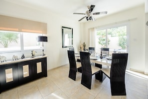 Un coin repas comprenant une console noire, quatre chaises, un ventilateur de plafond et de grandes fenêtres donnant sur un paysage verdoyant.