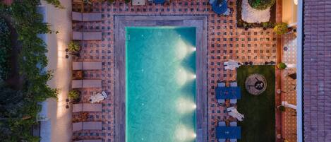 Luftaufnahme eines rechteckigen Schwimmbeckens mit einer gefliesten Sonnenterrasse, Liegestühlen und Tischen.