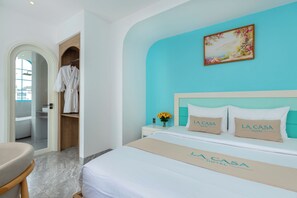 A hotel room with a bed, a nightstand, a vase with flowers, a painting on the wall, and a robe hanging on a door. - La Casa Hotel (Ho Chi Minh City)