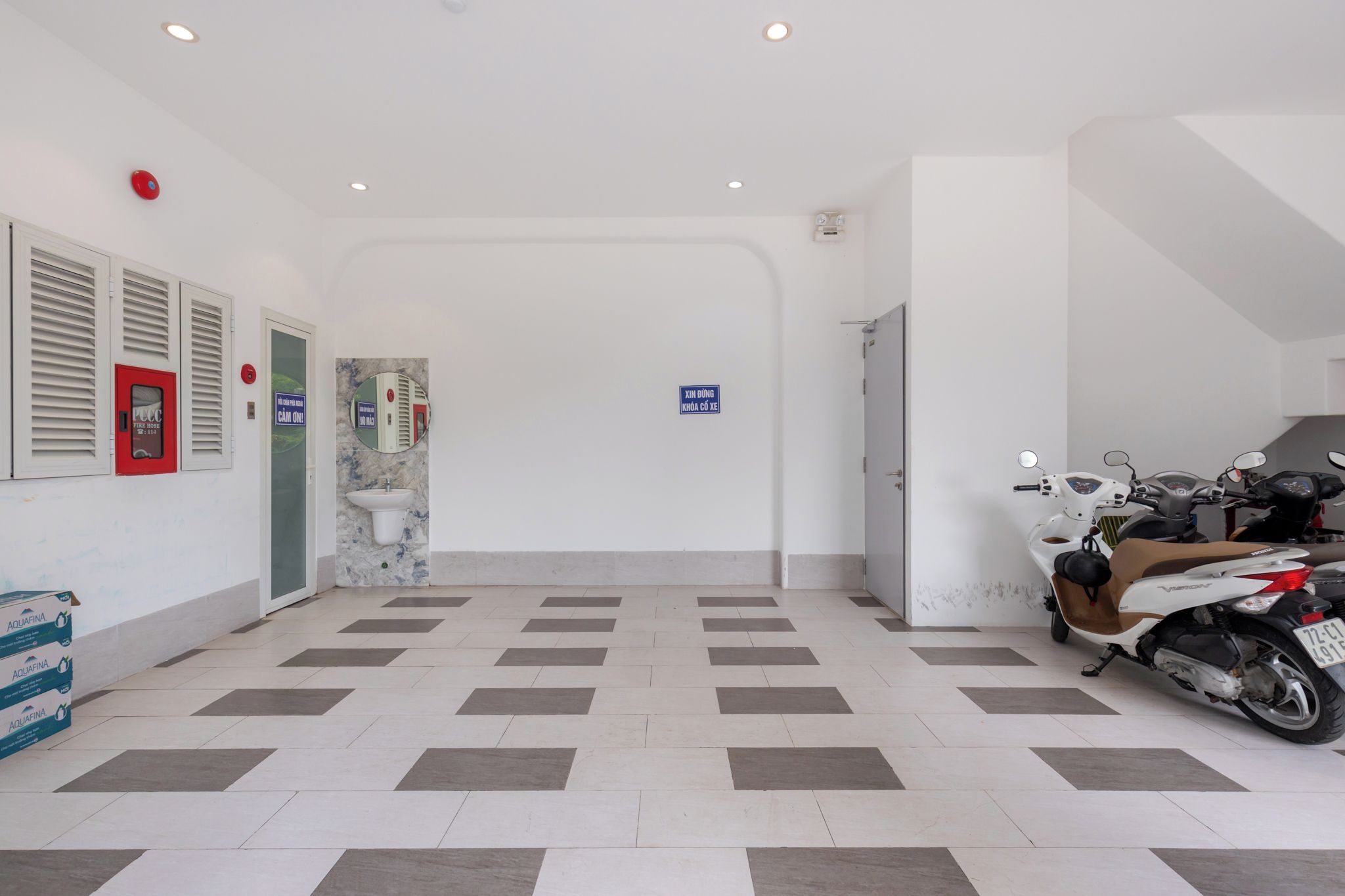 Un espacio interior limpio y bien iluminado, con suelo de baldosas, estación de manguera contra incendios, dispensador de agua y un scooter estacionado.