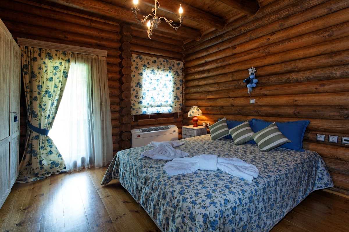 A log cabin room with a wooden bed, a window with floral curtains, a nightstand with a lamp, and a chandelier.