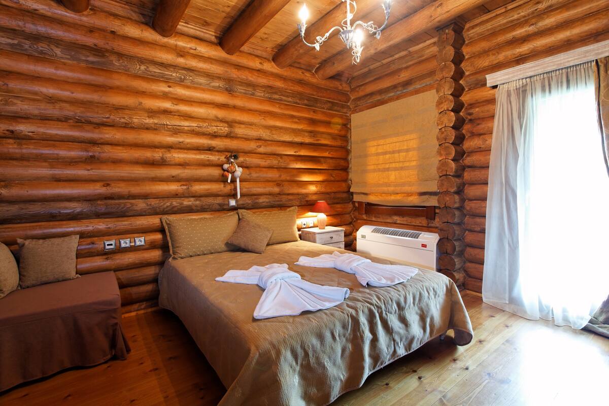 A log cabin bedroom with a wooden bed, a nightstand, a lamp, and a window with curtains.