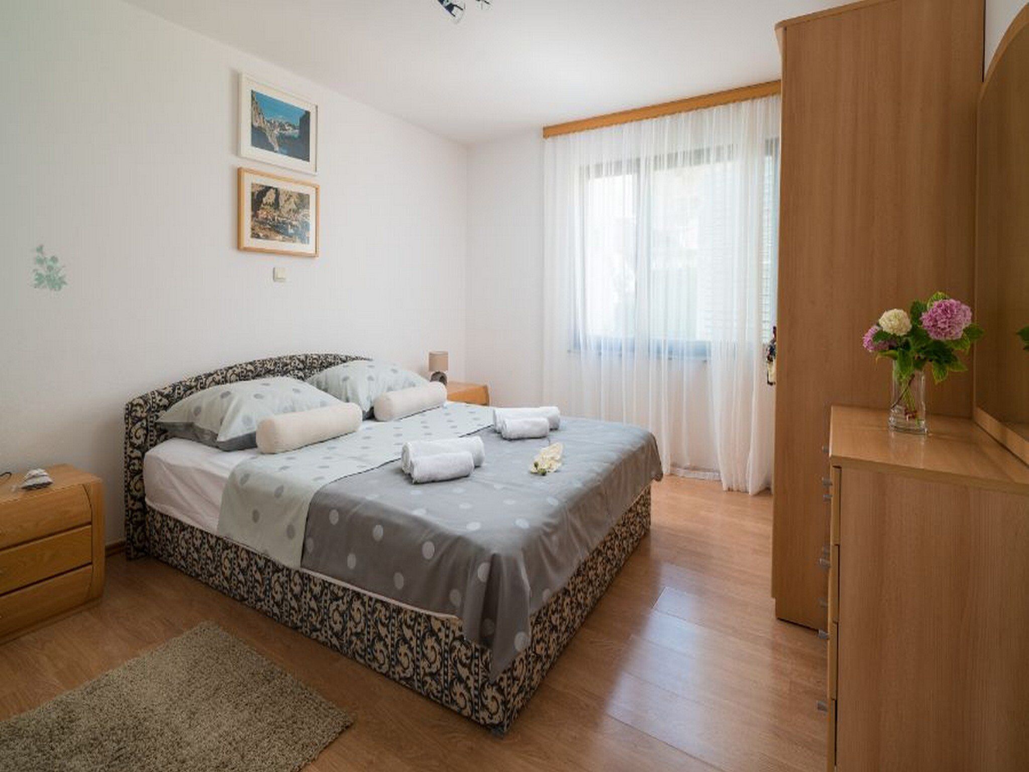 Ein ordentlich eingerichtetes Schlafzimmer mit Bett, Nachttischen, einem Schminktisch und einem Fenster mit durchscheinenden Vorhängen.
