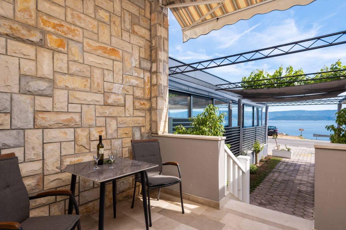 A stone-walled patio with a table and chairs, a glass enclosure with a view of the sea, and a covered outdoor area.