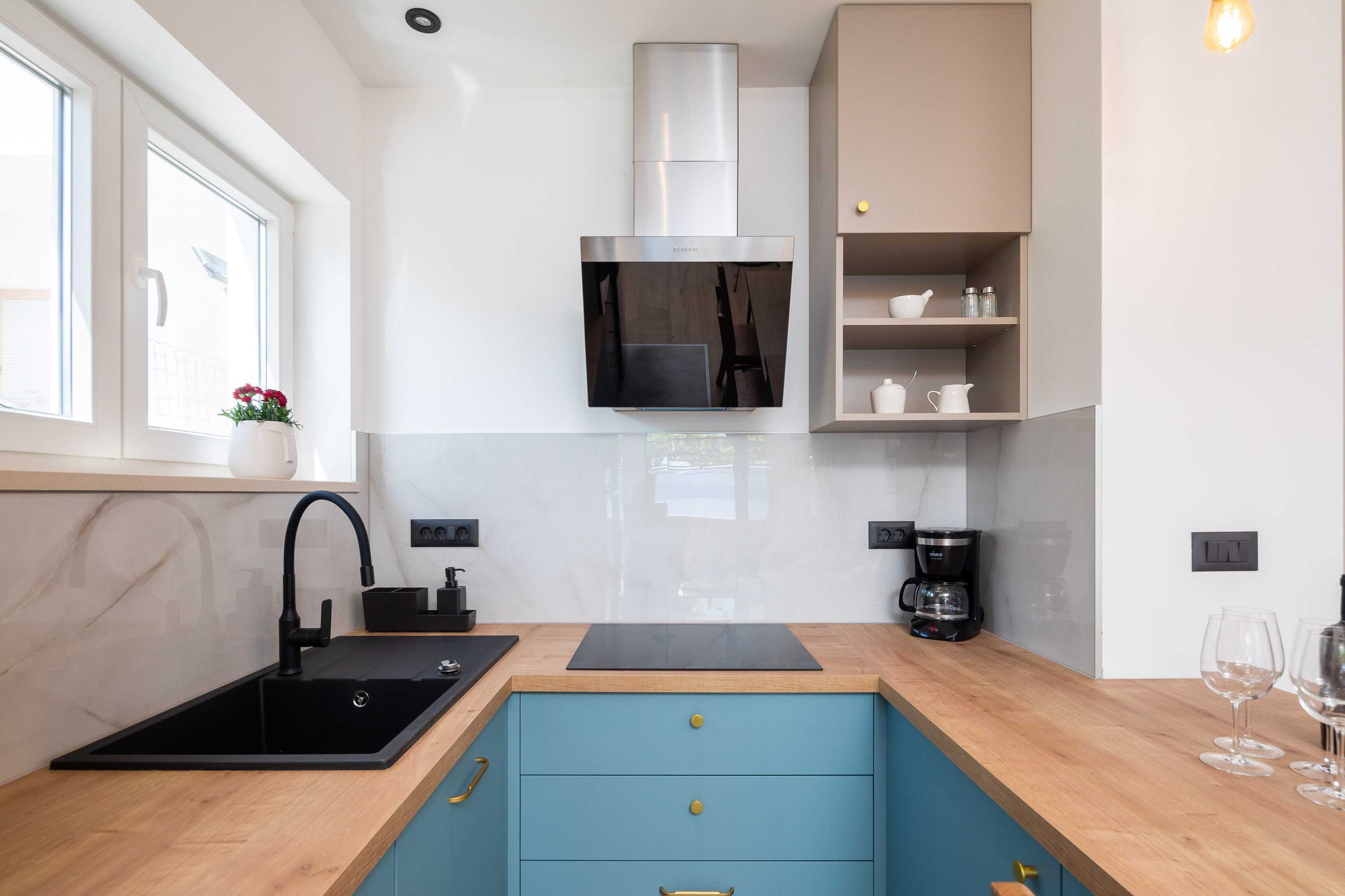 A modern kitchen with a black sink, blue cabinets, a stainless steel range hood, and a window with a flower vase.