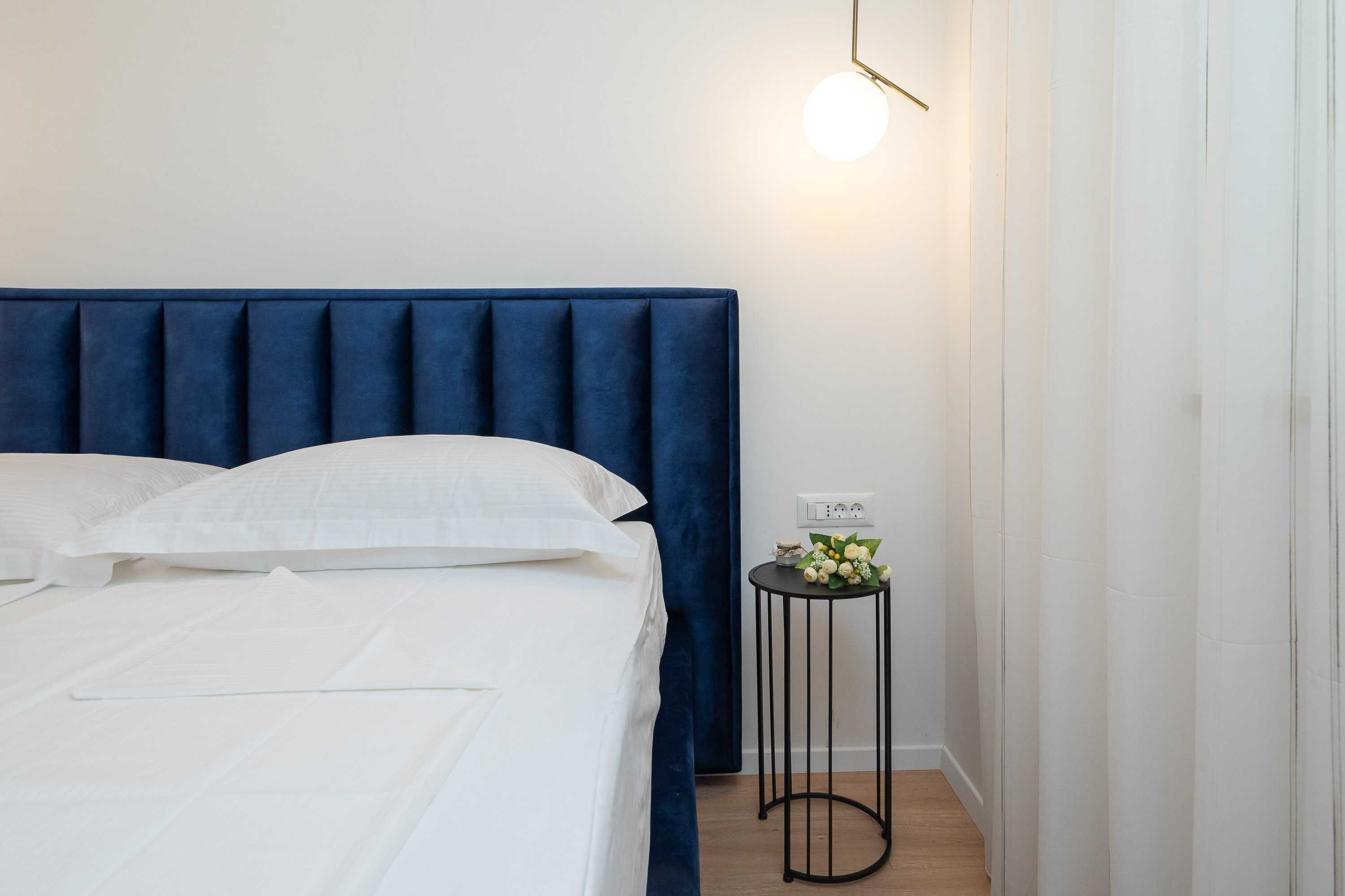 Un lit bien fait, avec du linge de lit blanc, une tête de lit rembourrée bleue et une petite table de chevet agrémentée d’un arrangement floral.