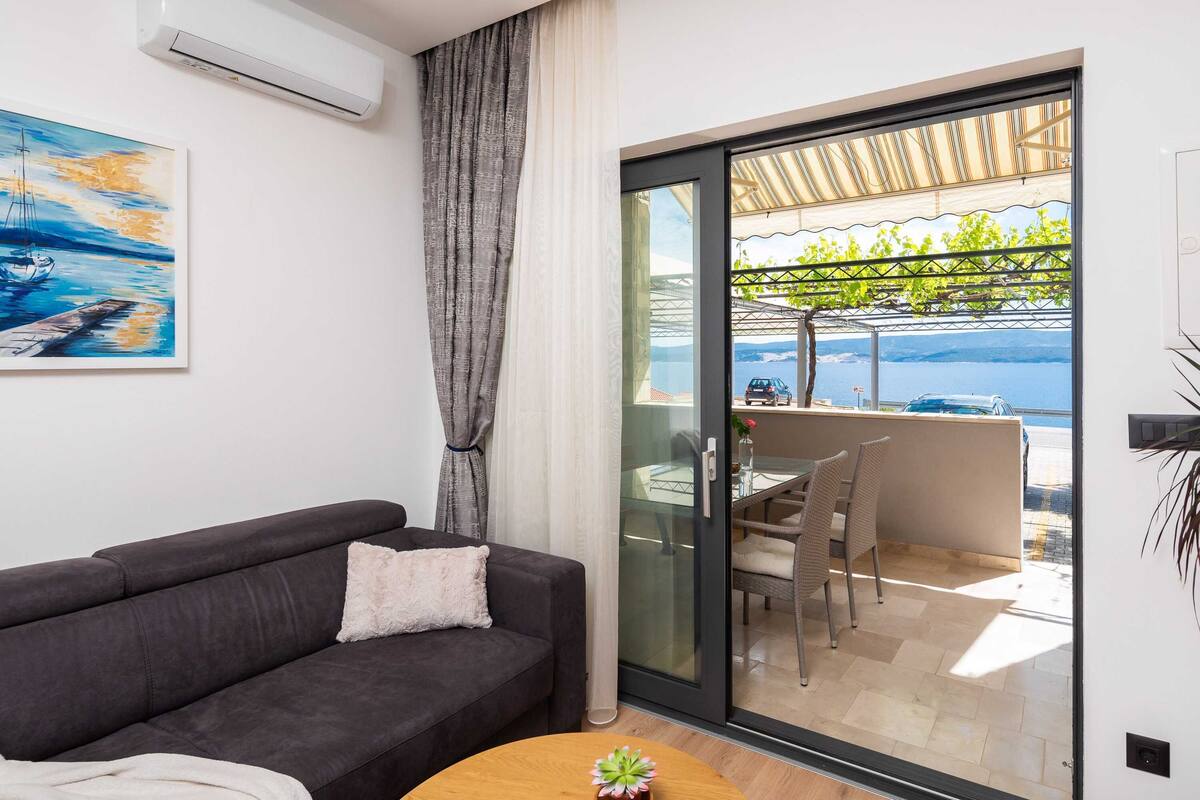 A modern living room with a dark sofa, a small round table, and a glass door leading to an outdoor area with a table and chairs.