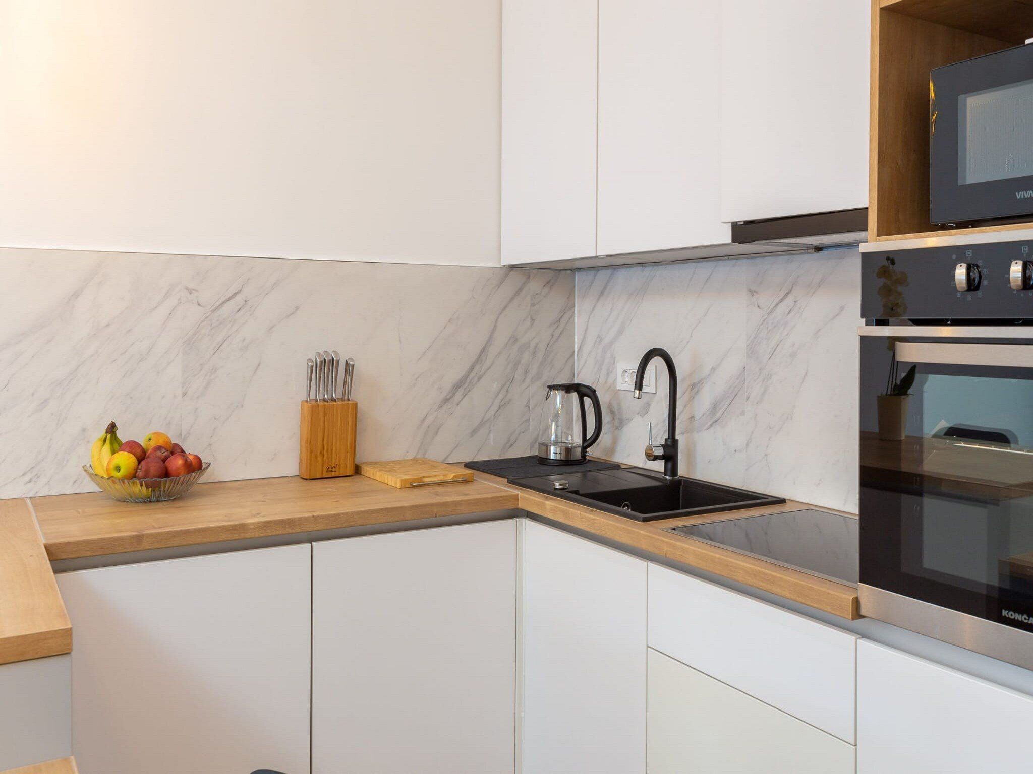 A modern kitchen with white cabinets, a black sink, a kettle, and a fruit bowl.