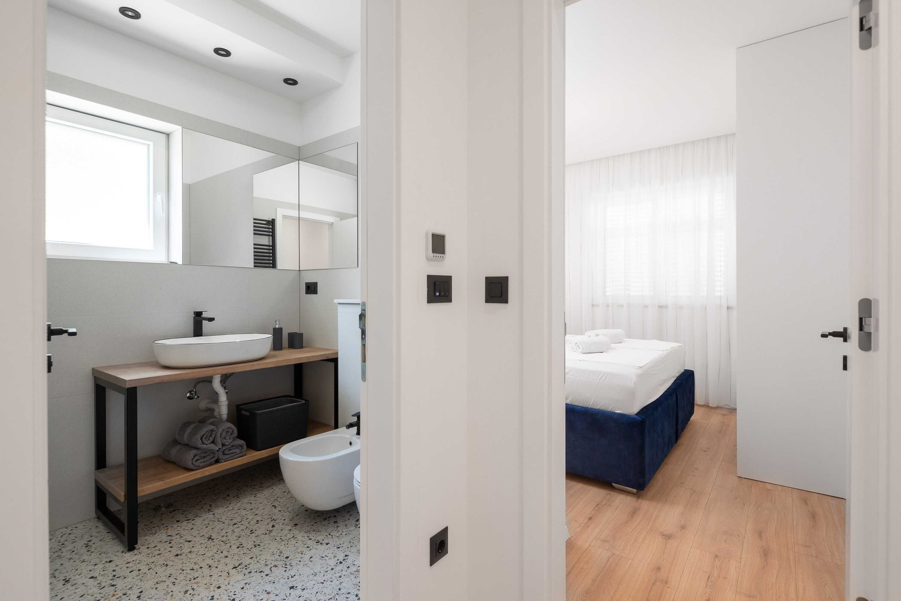 Un baño moderno con un espejo grande, un lavabo blanco y un bidé. El cuarto tiene piso de madera y una cama con cabecera azul en el dormitorio contiguo.