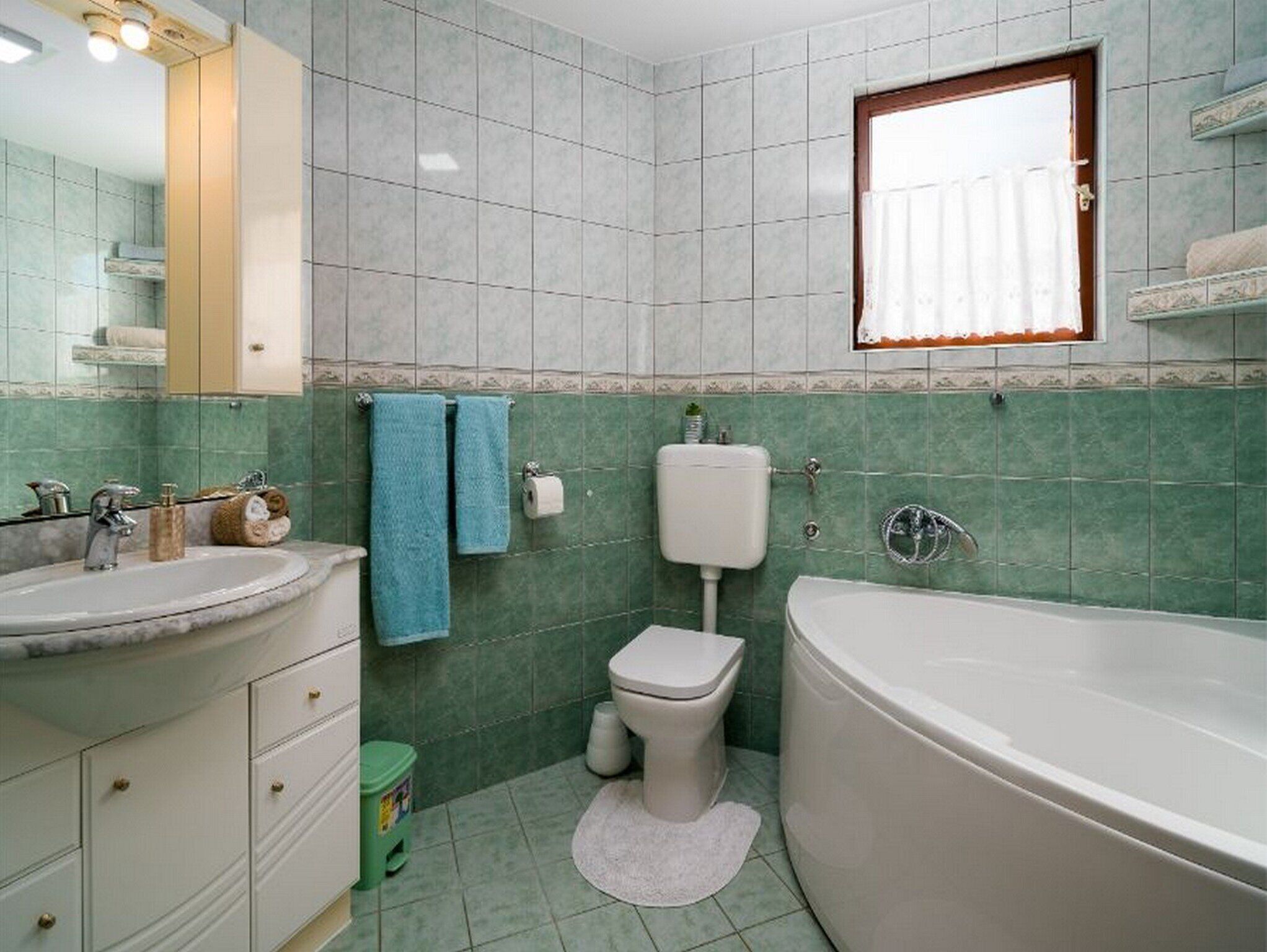 A bathroom with a sink, toilet, and bathtub, green tiled walls, and a window with curtains.