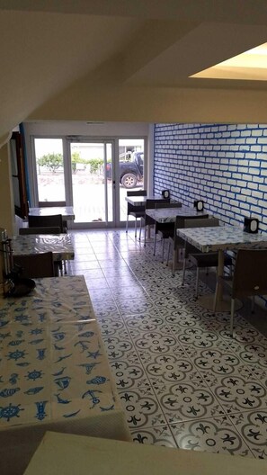 Uma área de refeições com ladrilhos estampados, mesas e cadeiras, e vista para o exterior através de uma porta de vidro.