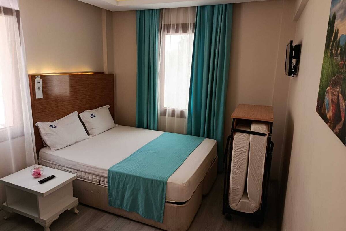 A hotel room with a bed, a bedside table, a nightstand, a window with curtains, and a painting on the wall.