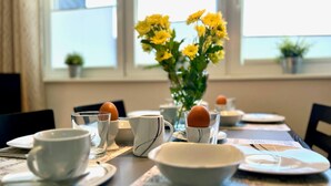A dining table set with white cups, bowls, and a glass with an egg. There is a vase with yellow flowers in the background.