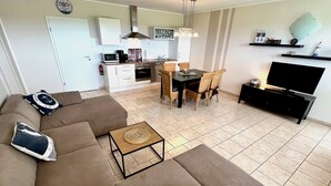 A modern living room with a sectional sofa, a dining area with a table and chairs, a kitchen with white cabinets, and a flat-screen TV on a stand. - Well-kept vacation complex with 5 residential units in a quiet cul-de-sac location (Dahme)