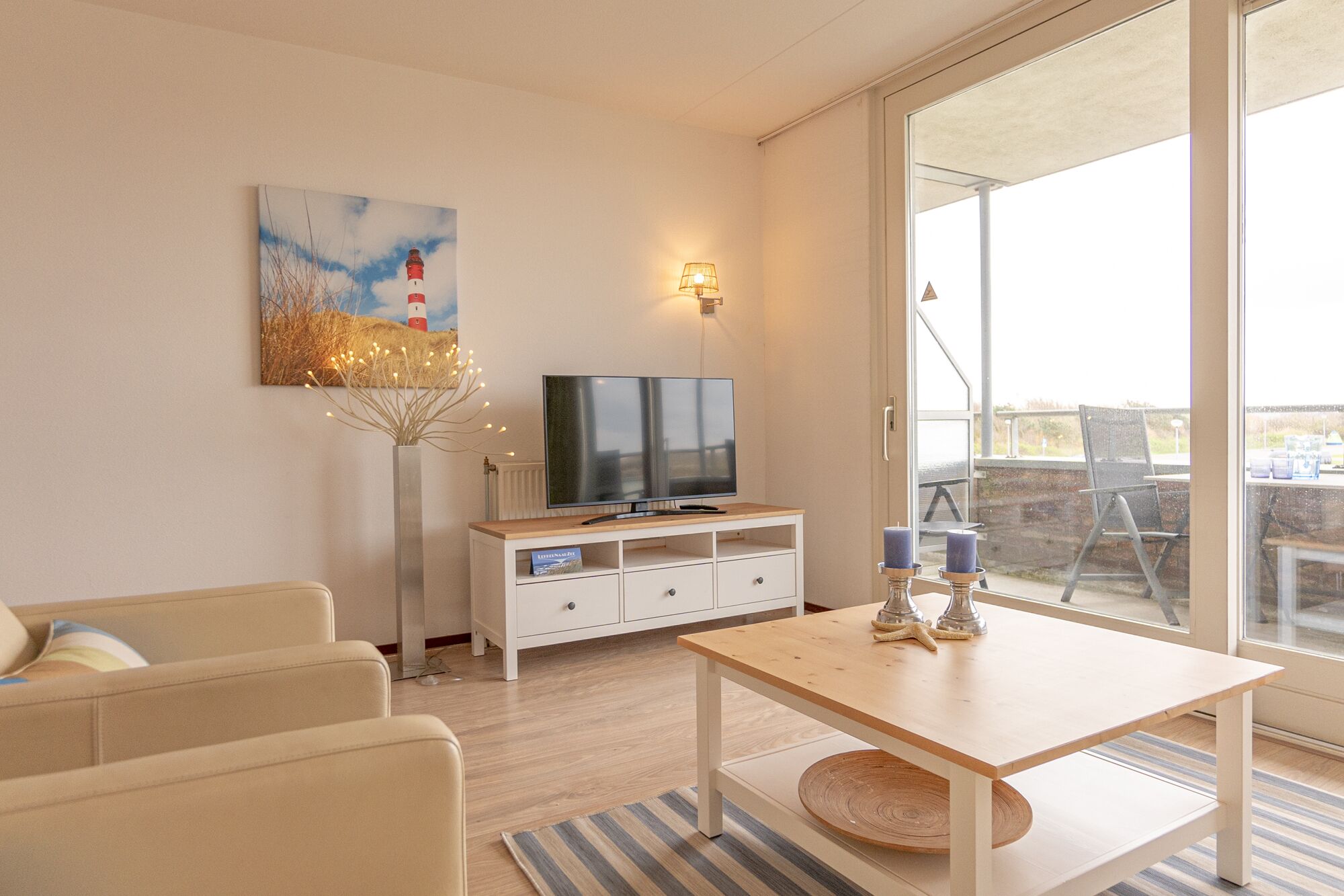Un salon moderne avec un téléviseur à écran plat posé sur une console blanche, un tableau représentant un phare et une grande porte-fenêtre coulissante donnant sur un espace extérieur.