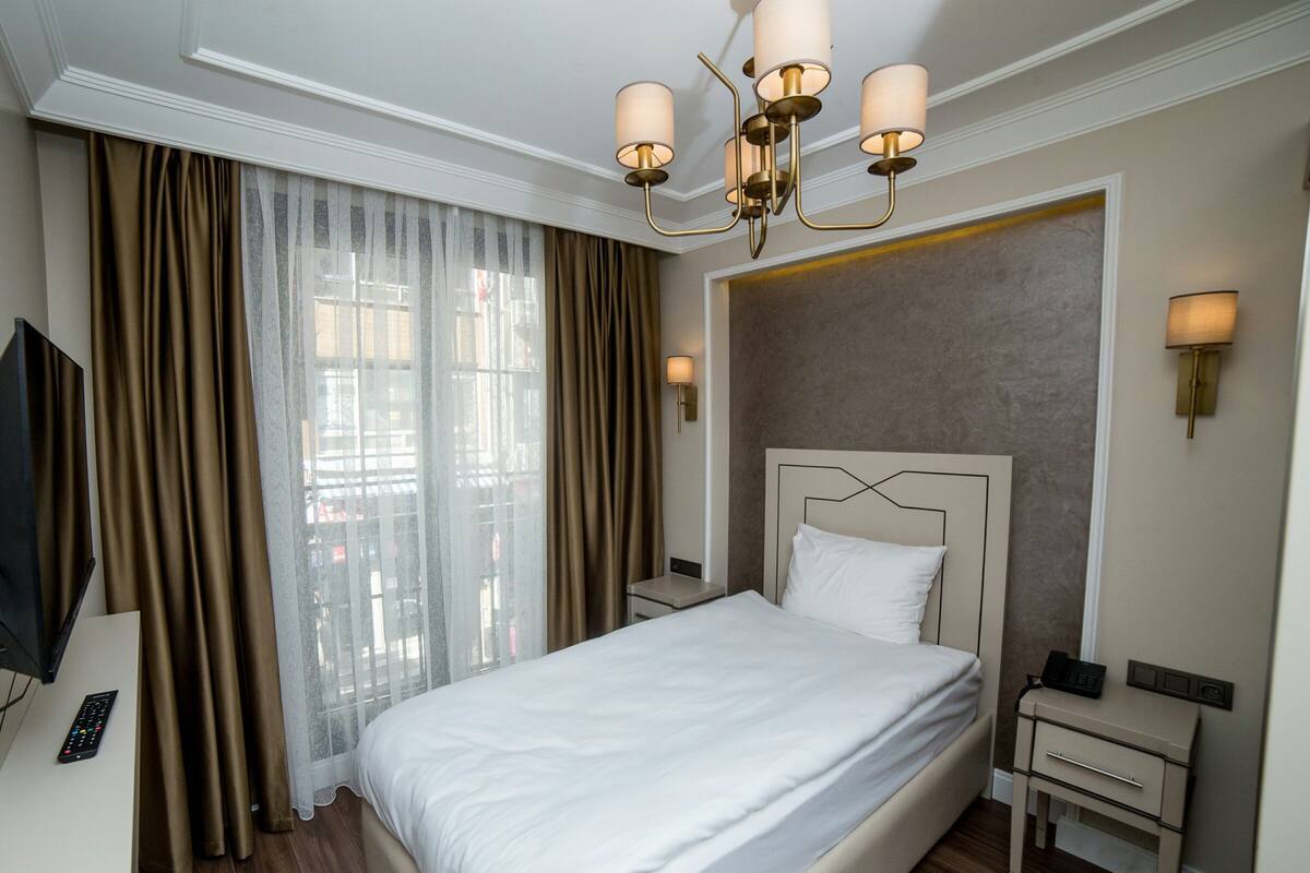 Une chambre d’hôtel avec un grand lit, une télévision, une table de chevet avec une lampe et une vue sur la ville à travers de fines rideaux.