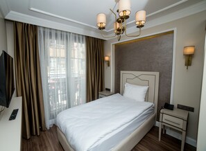 Een hotelkamer met een groot bed, een televisie, een nachtkastje met een lamp en uitzicht op de stad door dunne gordijnen.